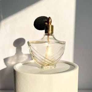 May include: A vintage perfume bottle with a clear, curved glass body and a gold-colored atomizer. The atomizer has a black, textured bulb. The bottle is displayed on a white, cylindrical stand, with shadows cast on a white background.