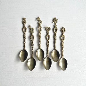 May include: Six antique brass spoons with decorative handles. Each spoon has a different design at the top, including a crown, deer, monkey, and floral motifs. The spoons are displayed on a white surface.