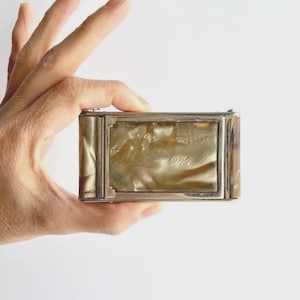 May include: A rectangular, vintage silver-toned box with a shimmering, pearlescent interior. The box has a hinged lid and is held in a hand against a white background. The box's design suggests it may be a jewelry or trinket box.