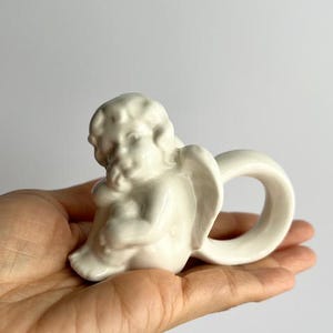 K&ouml;nnte beinhalten: Kleine, cremefarbene Keramik-Engelsfigur mit einem Ringgriff. Der Engel sitzt, die Fl&uuml;gel leicht ausgebreitet. Die Figur hat eine glatte, gl&auml;nzende Oberfl&auml;che.