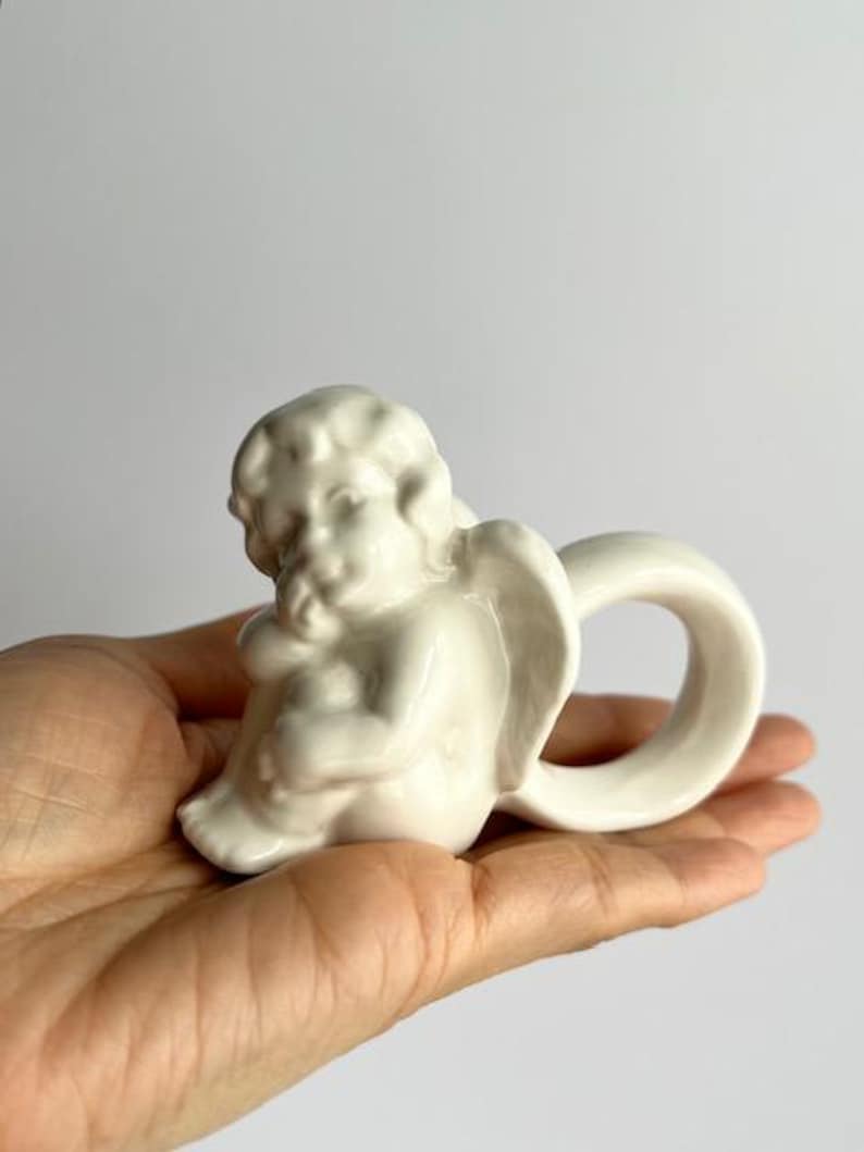 K&ouml;nnte beinhalten: Kleine, cremefarbene Keramik-Engelsfigur mit einem Ringgriff. Der Engel sitzt, die Fl&uuml;gel leicht ausgebreitet. Die Figur hat eine glatte, gl&auml;nzende Oberfl&auml;che.