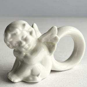 K&ouml;nnte beinhalten: Ein wei&szlig;er Serviettenring aus Keramik in Form eines Cherubs. Der Cherub sitzt mit ausgebreiteten Fl&uuml;geln, und der Ring wird durch einen gebogenen Abschnitt gebildet. Das St&uuml;ck hat eine gl&auml;nzende Oberfl&auml;che und ist f&uuml;r die Tischdekoration bestimmt.