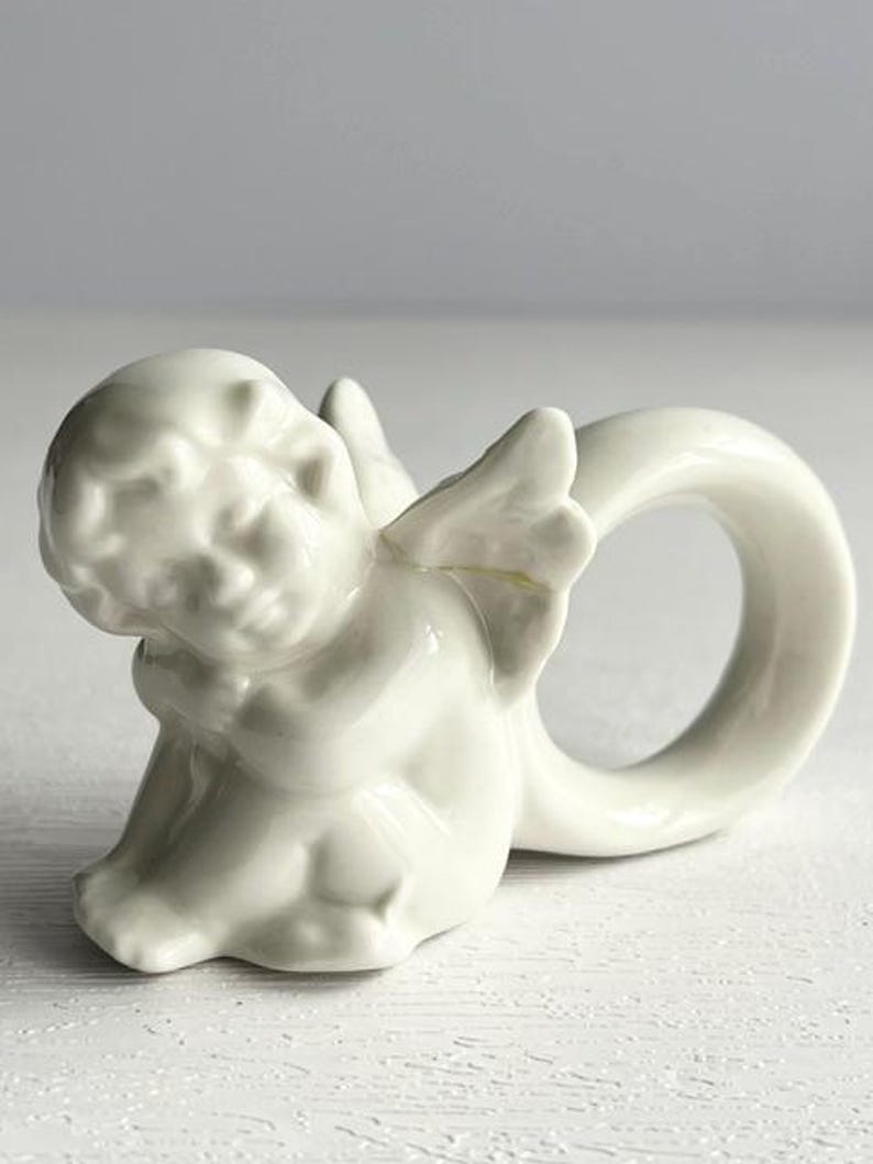 K&ouml;nnte beinhalten: Ein wei&szlig;er Serviettenring aus Keramik in Form eines Cherubs. Der Cherub sitzt mit ausgebreiteten Fl&uuml;geln, und der Ring wird durch einen gebogenen Abschnitt gebildet. Das St&uuml;ck hat eine gl&auml;nzende Oberfl&auml;che und ist f&uuml;r die Tischdekoration bestimmt.