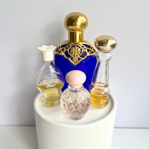 May include: A collection of vintage perfume bottles with gold and blue accents. The bottles vary in size and shape, with a variety of decorative stoppers. One bottle has the text "Here's my Heart Cologne". The bottles are displayed on a white pedestal.