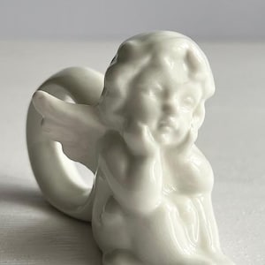 K&ouml;nnte beinhalten: Ein wei&szlig;er Serviettenring in Form eines Engels aus Keramik. Der Engel ist in sitzender Position dargestellt, den Kopf auf die Hand gest&uuml;tzt. Die Fl&uuml;gel sind ausgebreitet, und der Ring befindet sich hinter dem Engel. Das St&uuml;ck hat eine gl&auml;nzende Oberfl&auml;che.