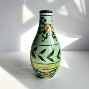 May include: A hand-painted ceramic vase with a light green base, adorned with floral and leaf motifs in red, yellow, and dark green. The vase has a rounded body and a narrow neck, perfect for displaying flowers. The vase is approximately 8 inches tall.