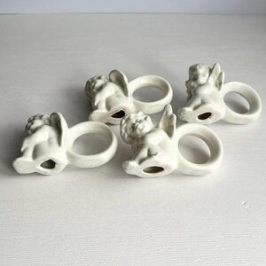 K&ouml;nnte beinhalten: Vier cremefarbene Serviettenringe aus Keramik, jeweils mit einem Cherub-Design. Die Cherubim haben Fl&uuml;gel und sitzen, wobei der Ring die Basis bildet. Die Serviettenringe sind auf einer wei&szlig;en Oberfl&auml;che angeordnet.