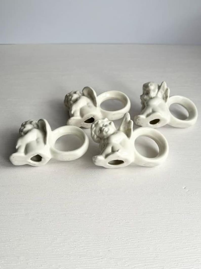 K&ouml;nnte beinhalten: Vier cremefarbene Serviettenringe aus Keramik, jeweils mit einem Cherub-Design. Die Cherubim haben Fl&uuml;gel und sitzen, wobei der Ring die Basis bildet. Die Serviettenringe sind auf einer wei&szlig;en Oberfl&auml;che angeordnet.