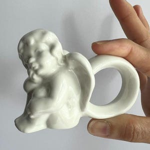 K&ouml;nnte beinhalten: Ein wei&szlig;er Keramik-Serviettenring mit einem Cherub-Design. Der Cherub ist in sitzender Pose mit gefalteten Fl&uuml;geln dargestellt, und der Ring hat eine einfache, abgerundete Form. Der Stil ist klassisch und dekorativ.