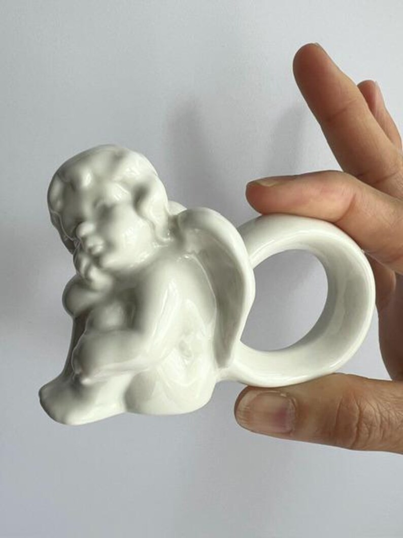 K&ouml;nnte beinhalten: Ein wei&szlig;er Keramik-Serviettenring mit einem Cherub-Design. Der Cherub ist in sitzender Pose mit gefalteten Fl&uuml;geln dargestellt, und der Ring hat eine einfache, abgerundete Form. Der Stil ist klassisch und dekorativ.