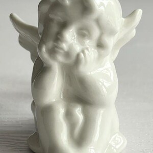 K&ouml;nnte beinhalten: Kleine, wei&szlig;e Cherub-Figur. Der Cherub sitzt mit dem Kinn auf den H&auml;nden und hat kleine Fl&uuml;gel. Die Figur besteht aus einem gl&auml;nzenden, glatten Material und hat ein klassisches, engelhaftes Aussehen.