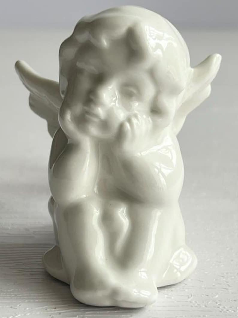 K&ouml;nnte beinhalten: Kleine, wei&szlig;e Cherub-Figur. Der Cherub sitzt mit dem Kinn auf den H&auml;nden und hat kleine Fl&uuml;gel. Die Figur besteht aus einem gl&auml;nzenden, glatten Material und hat ein klassisches, engelhaftes Aussehen.