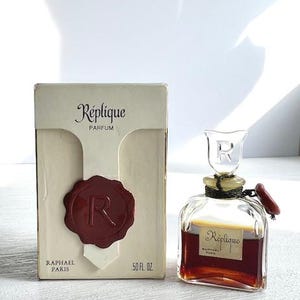 May include: A vintage perfume bottle and box set. The box is off-white with the words "Réplique PARFUM" and a red wax seal. The perfume bottle is square with a clear stopper and contains a dark amber liquid. The box reads "RAPHAEL PARIS" and "50 fl oz."
