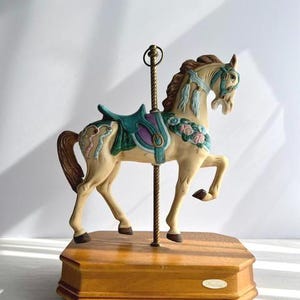 May include: A carousel horse figurine in cream, adorned with teal and purple saddle details, and floral accents. The horse stands on a wooden base with a brass pole and ring. The figurine is a decorative item.