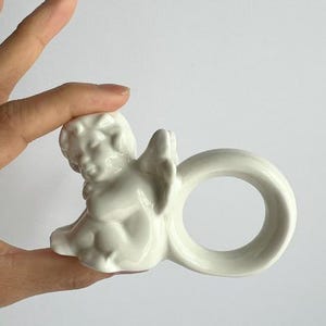 K&ouml;nnte beinhalten: Ein wei&szlig;er Serviettenring aus Keramik mit einem Cherub-Design. Der Cherub sitzt mit ausgebreiteten Fl&uuml;geln, und der Ring hat eine glatte, kreisf&ouml;rmige Form. Das St&uuml;ck ist wahrscheinlich f&uuml;r dekorative Tischdekorationen gedacht.