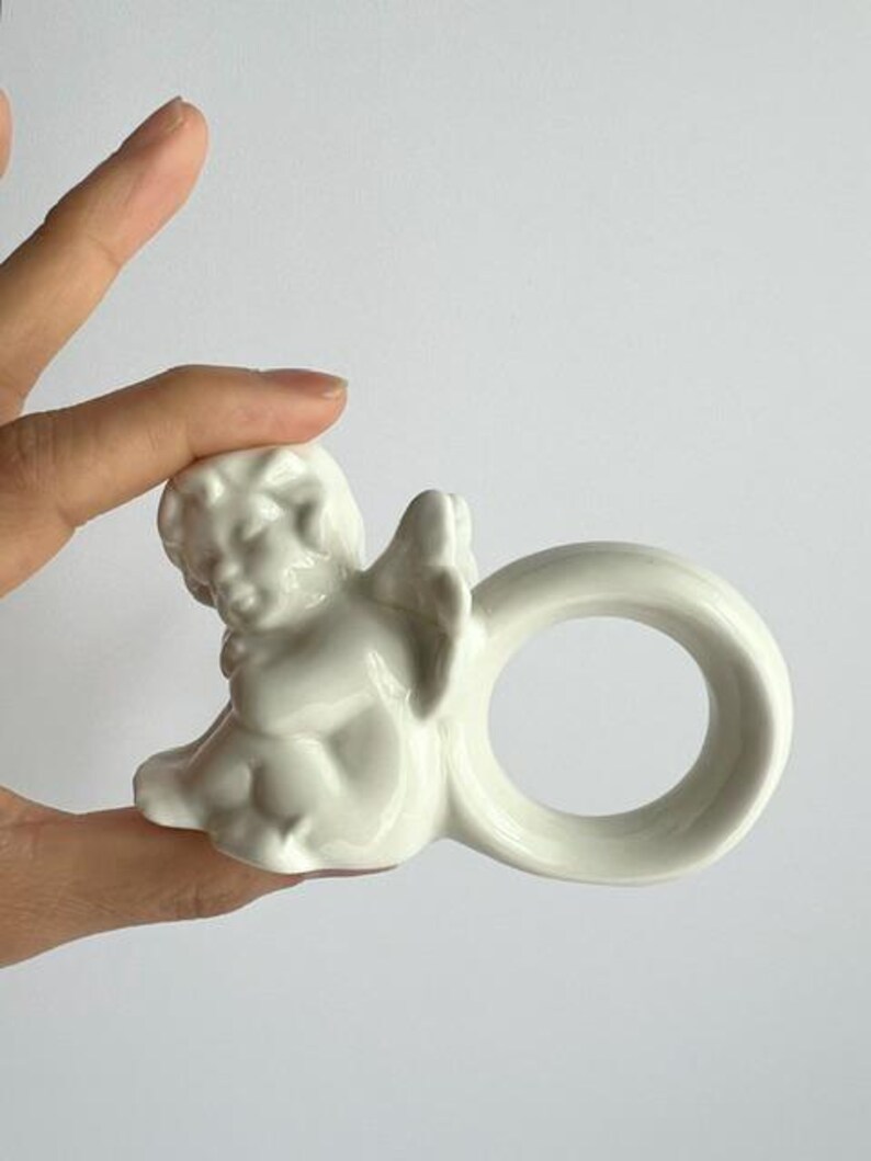 K&ouml;nnte beinhalten: Ein wei&szlig;er Serviettenring aus Keramik mit einem Cherub-Design. Der Cherub sitzt mit ausgebreiteten Fl&uuml;geln, und der Ring hat eine glatte, kreisf&ouml;rmige Form. Das St&uuml;ck ist wahrscheinlich f&uuml;r dekorative Tischdekorationen gedacht.