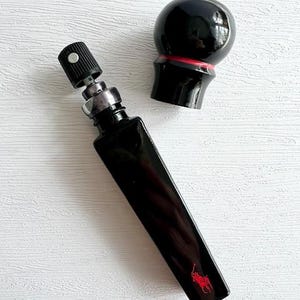 May include: A black rectangular perfume bottle with a red logo of a person on a horse, and a black cap. The bottle is next to a black spherical cap with a red band. The perfume bottle is on a white surface.