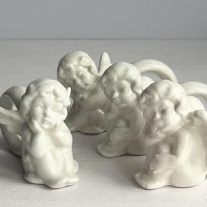K&ouml;nnte beinhalten: Vier wei&szlig;e Keramik-Puttenfiguren in verschiedenen Posen. Die Putten haben detaillierte Gesichtsz&uuml;ge und Fl&uuml;gel. Die Figuren sind auf einer wei&szlig;en Oberfl&auml;che angeordnet und schaffen eine ruhige und engelhafte Darstellung.