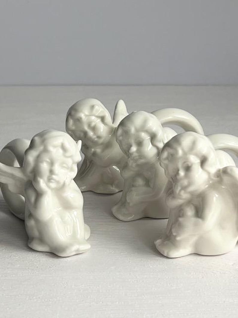 K&ouml;nnte beinhalten: Vier wei&szlig;e Keramik-Puttenfiguren in verschiedenen Posen. Die Putten haben detaillierte Gesichtsz&uuml;ge und Fl&uuml;gel. Die Figuren sind auf einer wei&szlig;en Oberfl&auml;che angeordnet und schaffen eine ruhige und engelhafte Darstellung.