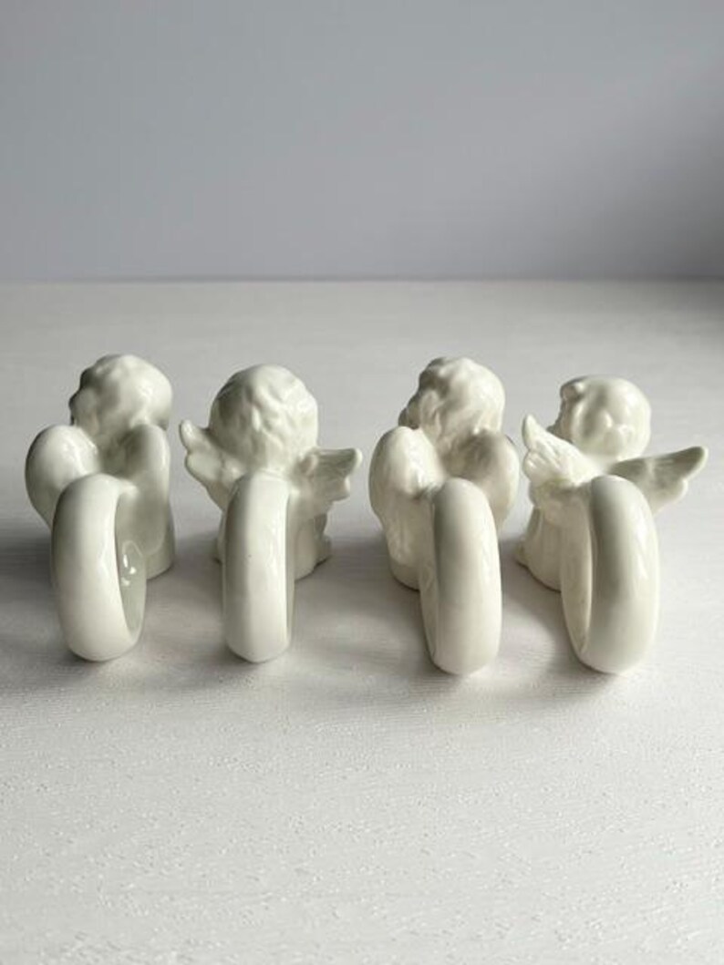 K&ouml;nnte beinhalten: Set mit vier wei&szlig;en Serviettenringen aus Keramik in Form von Engeln. Jede Engelsfigur hat detaillierte Fl&uuml;gel und eine Schlaufe zum Halten einer Serviette. Die Serviettenringe sind in einer Reihe angeordnet.