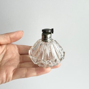 May include: A clear, faceted glass perfume bottle with a silver-colored metal cap. The bottle has a rounded, scalloped shape. The cap has a small, hinged top. The bottle is held in a hand against a white background.