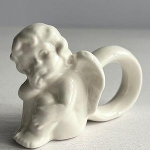 K&ouml;nnte beinhalten: Ein cremefarbener Serviettenring aus Keramik in Form eines Cherubs. Der Cherub sitzt mit ausgebreiteten Fl&uuml;geln, wobei ein Ring eine Schlaufe dahinter bildet. Das St&uuml;ck hat eine gl&auml;nzende Oberfl&auml;che und steht vor einem neutralen Hintergrund.