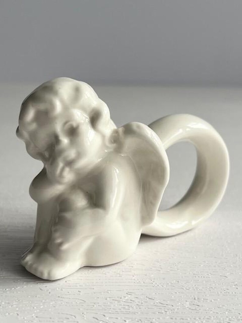 K&ouml;nnte beinhalten: Ein cremefarbener Serviettenring aus Keramik in Form eines Cherubs. Der Cherub sitzt mit ausgebreiteten Fl&uuml;geln, wobei ein Ring eine Schlaufe dahinter bildet. Das St&uuml;ck hat eine gl&auml;nzende Oberfl&auml;che und steht vor einem neutralen Hintergrund.