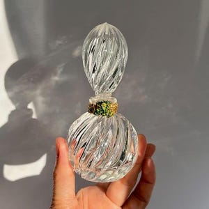 May include: A clear glass perfume bottle with a spiral design. The bottle has a round base and a long, pointed stopper. The neck of the bottle is decorated with green and gold embellishments. The bottle is held in a hand.