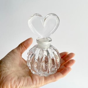 May include: Clear glass perfume bottle with a heart-shaped stopper. The bottle has a ribbed, rounded body and a clear, heart-shaped stopper. The bottle is held in a hand against a white background. A decorative item for a vanity or dresser.