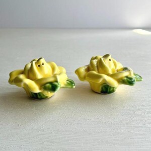May include: A pair of ceramic salt and pepper shakers shaped like blooming yellow roses. Each flower features bright yellow petals with green leaves at the base. The top of each rose has small holes for dispensing seasoning, crafted with a glossy finish.