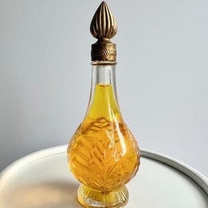 May include: A vintage perfume bottle with a gold-colored, ornate stopper. The clear glass bottle has a leaf design and is filled with a yellow liquid. The bottle is sitting on a white surface.
