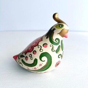 Peut inclure: Figurine d'oiseau en céramique avec une base crème, ornée de motifs décoratifs roses, verts et dorés. L'oiseau a une crête dorée, un bec rose et des accents verts et dorés autour des yeux. La queue est rouge.