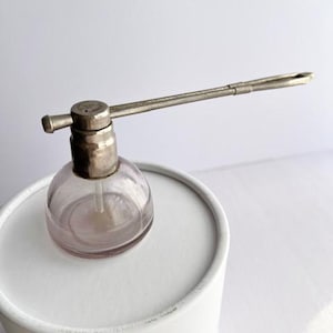 May include: A vintage perfume atomizer with a clear, round glass bottle and a silver-toned metal pump and spray arm. The bottle sits atop a white cylindrical container. The atomizer's spray arm extends horizontally.