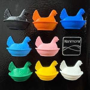 May include: A selection of fridge magnets featuring hen designs in baskets. The magnets are in a variety of colours: black, orange, blue, pink, green, yellow, and white. A Kenmore magnet is also present.