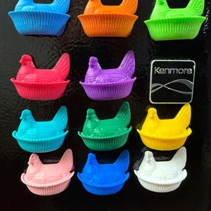 Pode incluir: Uma coleção de ímanes coloridos, impressos em 3D, em forma de galinha. Os ímanes são de várias cores, incluindo azul, laranja, verde, vermelho, roxo, turquesa, rosa, amarelo e branco. Cada galinha está numa base em forma de cesta. Um íman de frigorífico Kenmore também é visível.