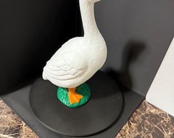 May include: A white goose figurine with an orange beak and feet, standing on a green base. The goose is on a black circular base.