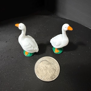 May include: Two white goose figurines with orange beaks and green feet. The figurines are standing on a black surface.