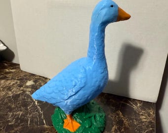 May include: A blue goose figurine with an orange beak and feet, standing on a green base with orange accents. The goose is a garden decoration.
