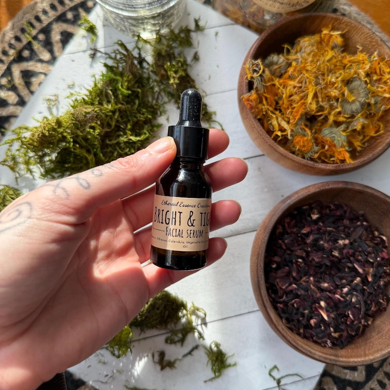 Może przedstawiać: Mała brązowa szklana butelka "BRIGHT & TIG - FACIAL SERUM" z czarną pipetą, trzymana w dłoni. Butelka otoczona jest suszonymi ziołami w drewnianych miskach i zielonym mchem. Serum jest oznaczone białym tekstem.