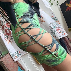Könnte beinhalten: Ein grüner und schwarzer Minirock mit Batikmuster und einem einzigartigen Ausschnitt-Design. Der Rock hat kreuzweise Schnürungen an den Seiten, die ein unverwechselbares visuelles Element hinzufügen. Der Stoff hat ein lebendiges, auffälliges Muster.