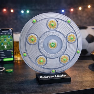 Op de afbeelding: Een cirkelvormige zilverkleurige trofee met de in reliëf aangebrachte tekst "DEUTSCHER FUSSBALLMEISTER" en groene accenten, gemonteerd op een zwarte rechthoekige voet met de witte tekst "Kickbase Meister" op de voorzijde als sportprijs.