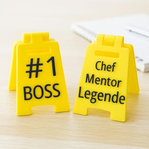 Puede incluir: Dos letreros amarillos plegables con texto negro. Un letrero dice "#1 BOSS", el otro dice "Chef Mentor Legende". Los letreros están sobre una superficie de madera de color claro, con un cuaderno y un bolígrafo en el fondo.