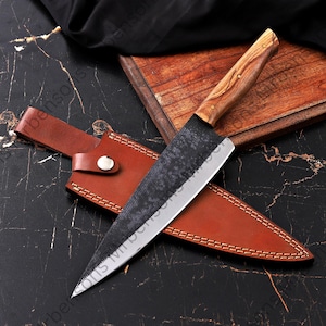 May include: A handmade chef's knife with a wooden handle and a textured, dark blade. The knife is displayed with a brown leather sheath, set against a dark, patterned surface. The blade is approximately 10 inches long.