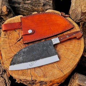 May include: A handmade chef's knife with a dark, textured blade and a wooden handle. The knife rests on a wooden surface next to a brown leather sheath with a strap and button closure. The blade has a polished edge.
