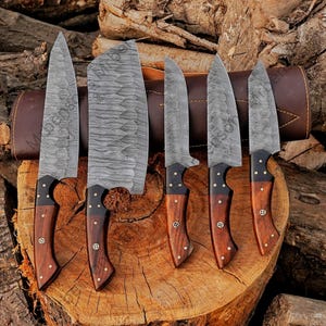 May include: A set of six Damascus steel knives with wooden handles. The blades have a unique patterned design. The handles are dark wood with brass rivets. A brown leather sheath is visible in the background.