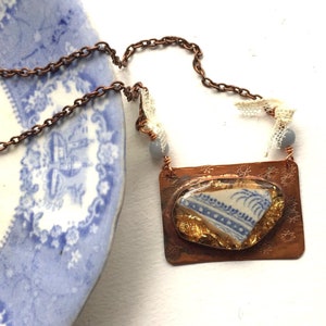 May include: A copper necklace with a blue and white porcelain shard pendant. The pendant is set in a copper frame and has gold leaf accents. The necklace is on a white background.