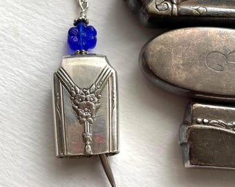 May include: A silver pendant necklace with a rectangular, Art Deco-style charm. The charm features floral detailing and a small pin extending from the bottom. A cluster of blue beads sits above the charm, connected to a silver chain.