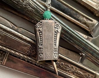 May include: A silver pendant necklace featuring a repurposed vintage utensil. The pendant has floral and beaded details, with a small green bead accent. The necklace is displayed against a backdrop of antique silverware.
