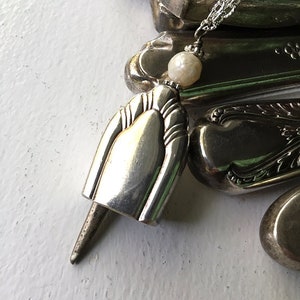 May include: A silver spoon pendant necklace with a pearl accent. The spoon is shaped like a bell and has a pointed end. The necklace is made from recycled silverware.