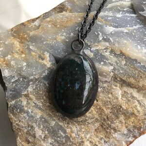 May include: Oval pendant necklace with a dark, mottled green stone set in a silver-tone metal frame. The pendant hangs from a delicate, dark chain. The stone has a polished, reflective surface, and the necklace rests on a textured, light-colored rock.